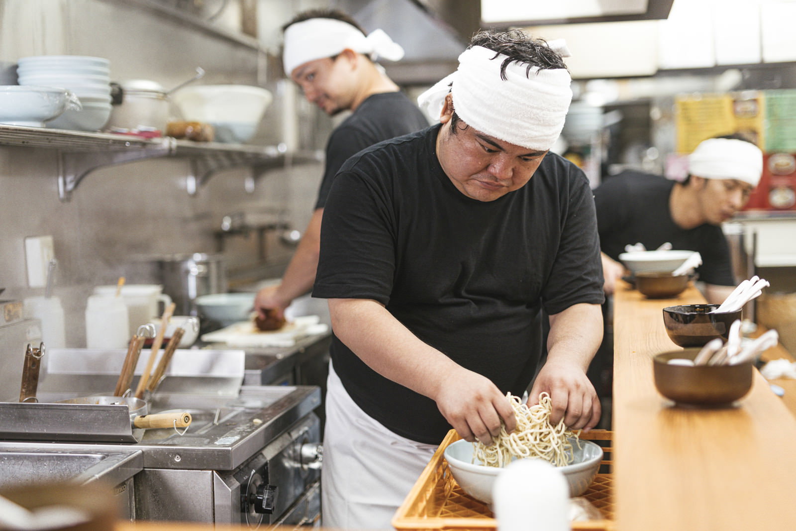 白い帽子とエプロンを身に着けた男性が厨房で茹でた麺をほぐしている［モデル：段田隼人］