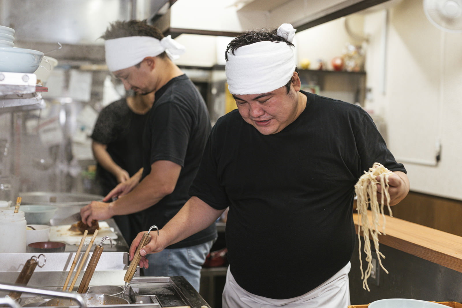 白い帽子とエプロンを着用した男性がラーメン店の厨房で麺を手に持ちテボをチェックしている［モデル：段田隼人 Max_Ezaki］