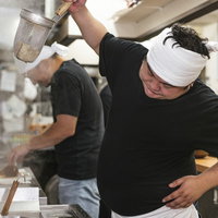 高い位置から湯切りにチャレンジするラーメン店主の厨房での一場面の写真