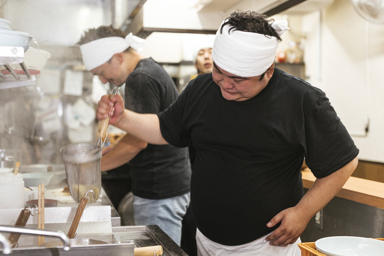 頭にタオルを巻いた男性が厨房でラーメンの湯切りをする［モデル：段田隼人 Max_Ezaki 大川竜弥］