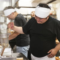白いタオルを頭に巻いて力強く湯切りするラーメン店主の男性の写真