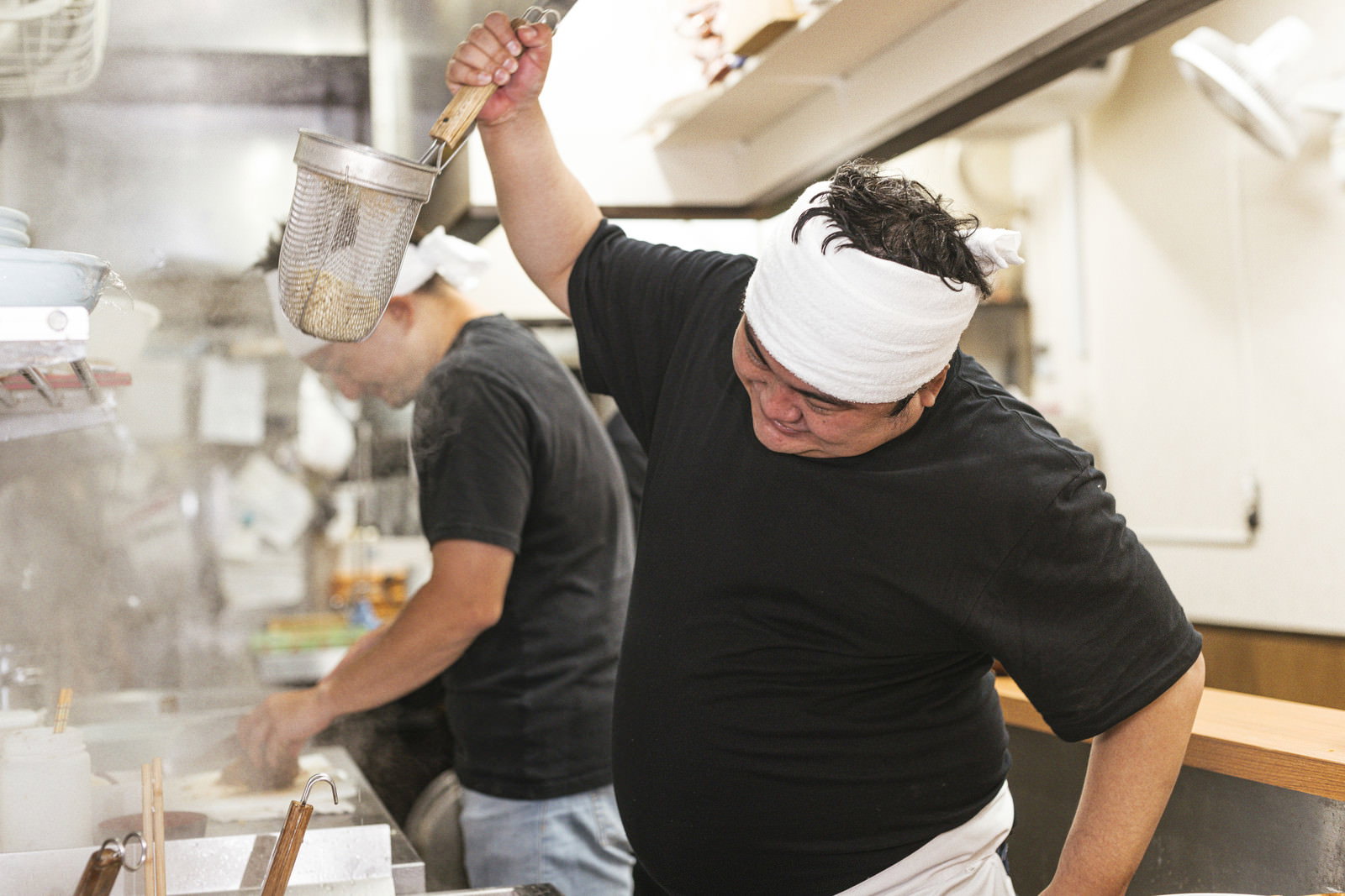 白い帽子とエプロン姿でテボを高く上げて湯切りするラーメン店主［モデル：段田隼人 Max_Ezaki］