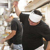 手を上げて勢いよく湯切りするラーメン店主の厨房作業の写真