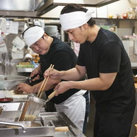 ラーメン店主にテボを使った麺茹でを教わる弟子の写真