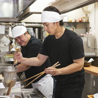 テボを使ってラーメンの湯切りにチャレンジする新人店員の写真