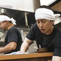 厨房から身を乗り出してカウンター席を拭くラーメン店のアルバイト男性の写真