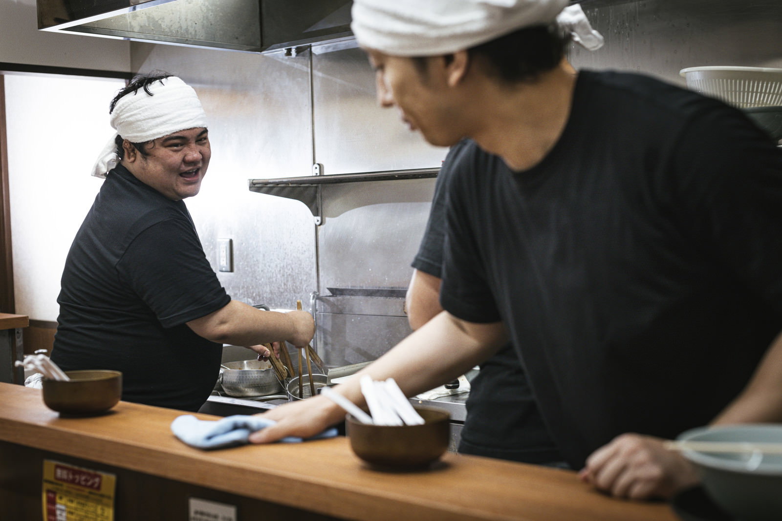 白い帽子とエプロンを着用したラーメン店の店主が厨房で調理作業をしている［モデル：段田隼人 大川竜弥］