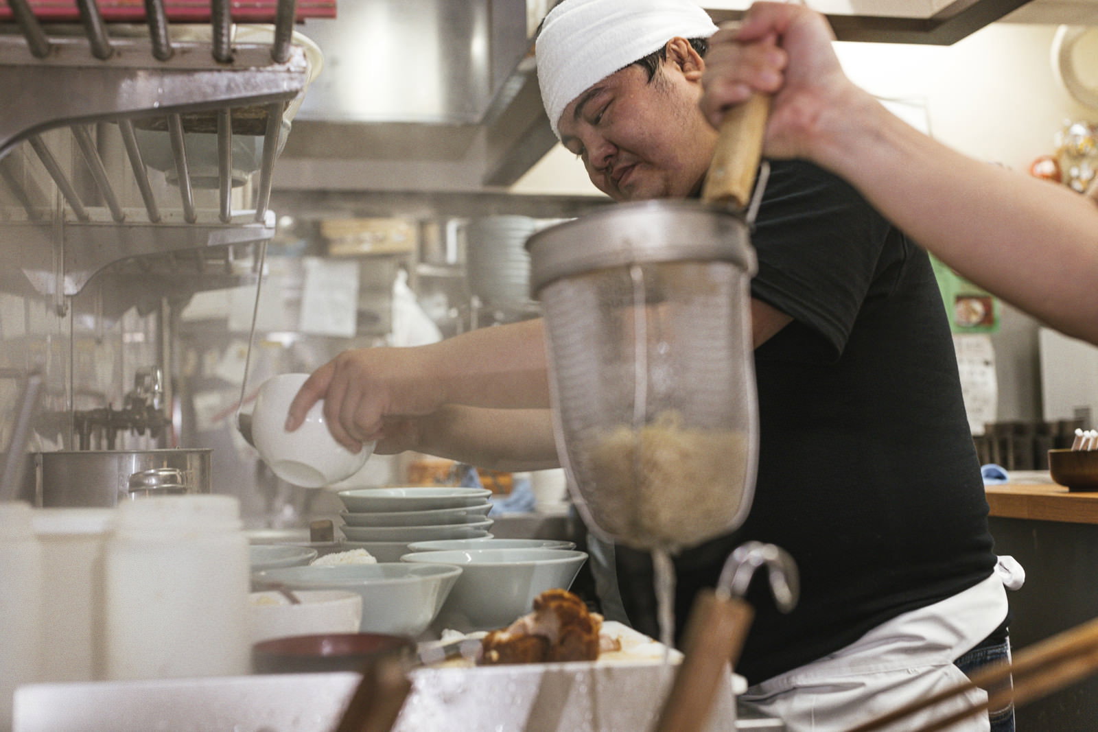 白い帽子をかぶったラーメン店主が厨房でテボを使って湯切りをしている［モデル：段田隼人］