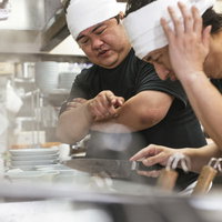 チャーシューの切り方指導で頭を下げる新人と師匠の写真