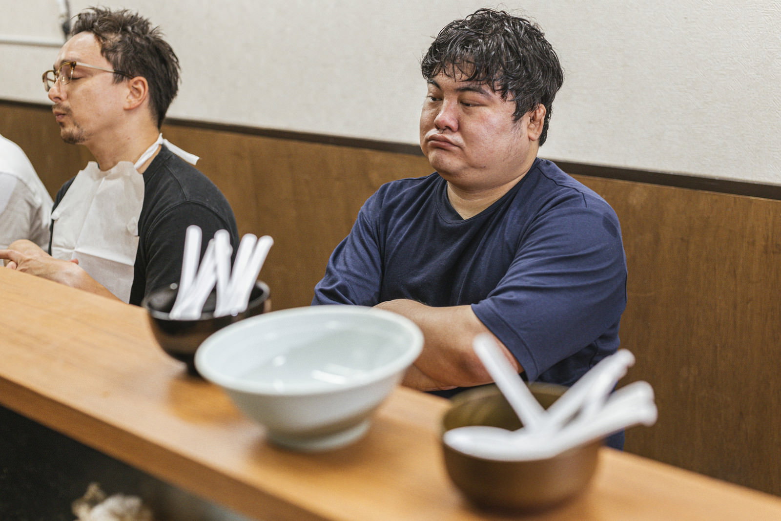 ラーメン店のカウンター席で壁の文字を見て頷く紺色Tシャツの男性［モデル：Max_Ezaki 段田隼人］