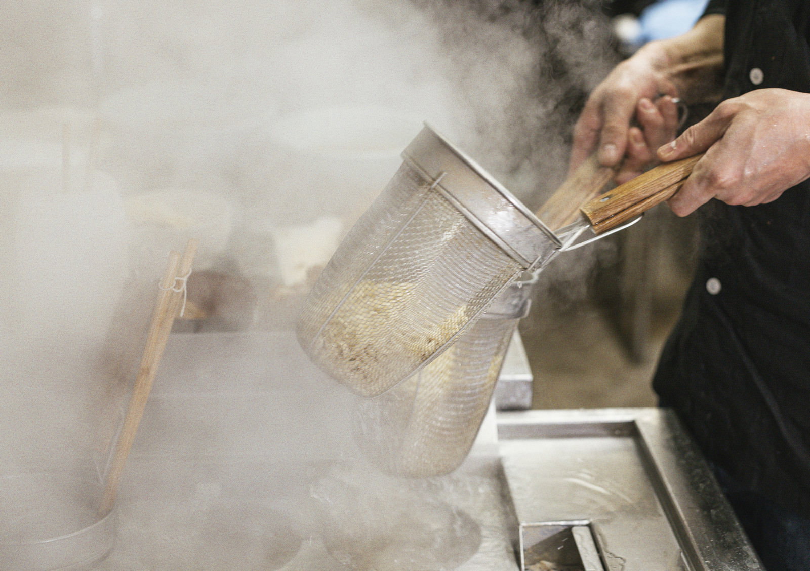 テボで麺の湯切りをする男性の手元、白い蒸気が立ち上る