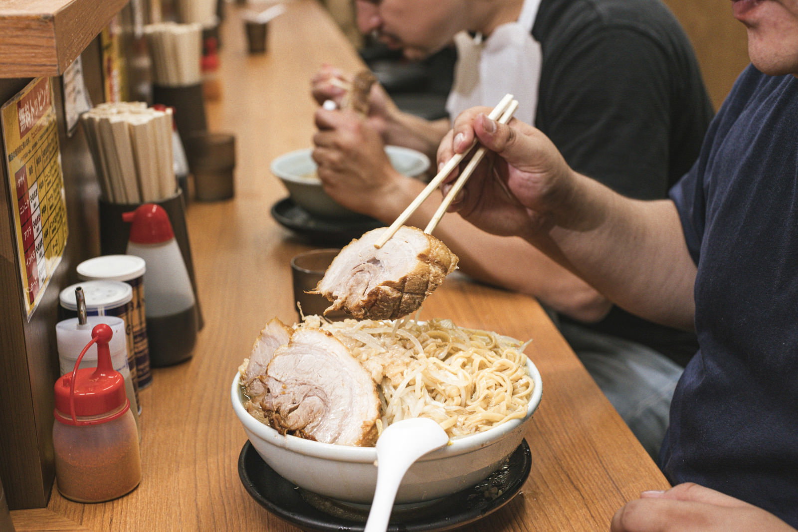 ラーメン店のカウンター席で分厚いチャーシューを箸で持ち上げて食べる男性［モデル：段田隼人］