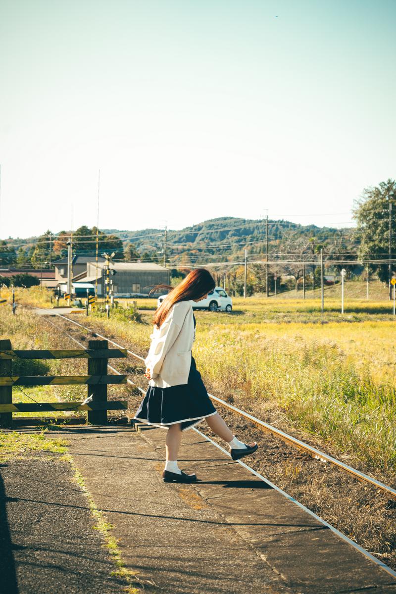 白いカーディガンを着た女性が田舎の駅のホームに立ち、背後に田園と山々がある風景［モデル：ラミエル］