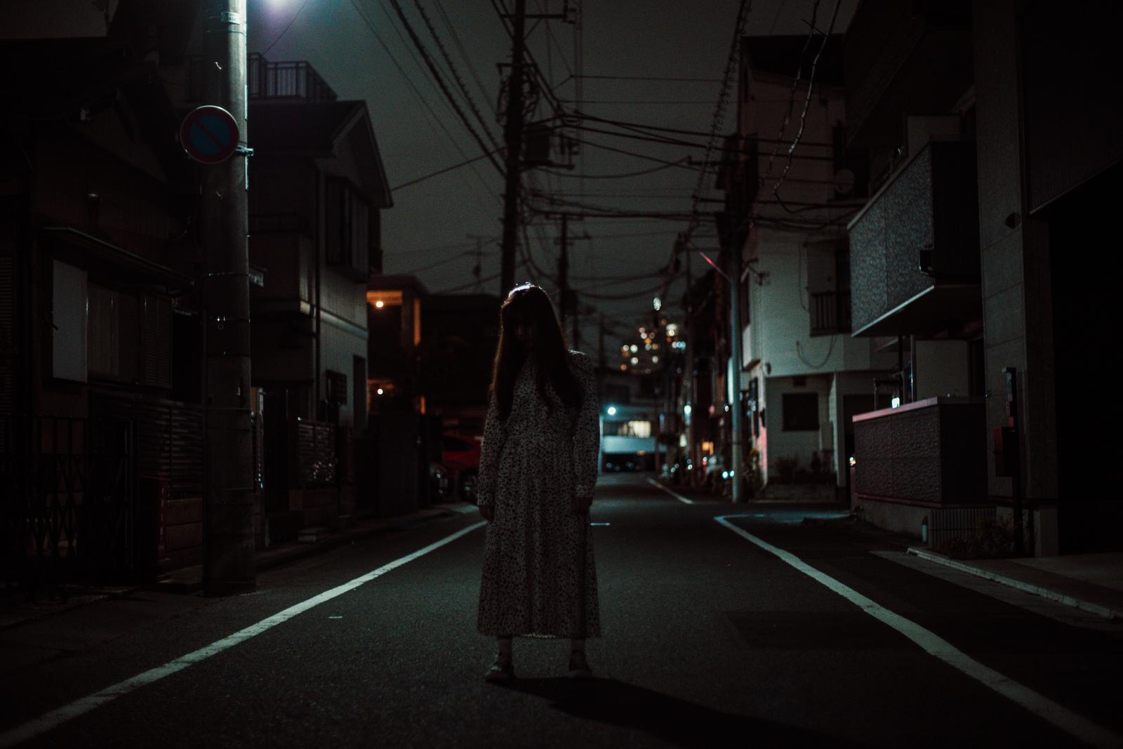 夜間の住宅街の中央に立つワンピース姿の女性［モデル：ラミエル］