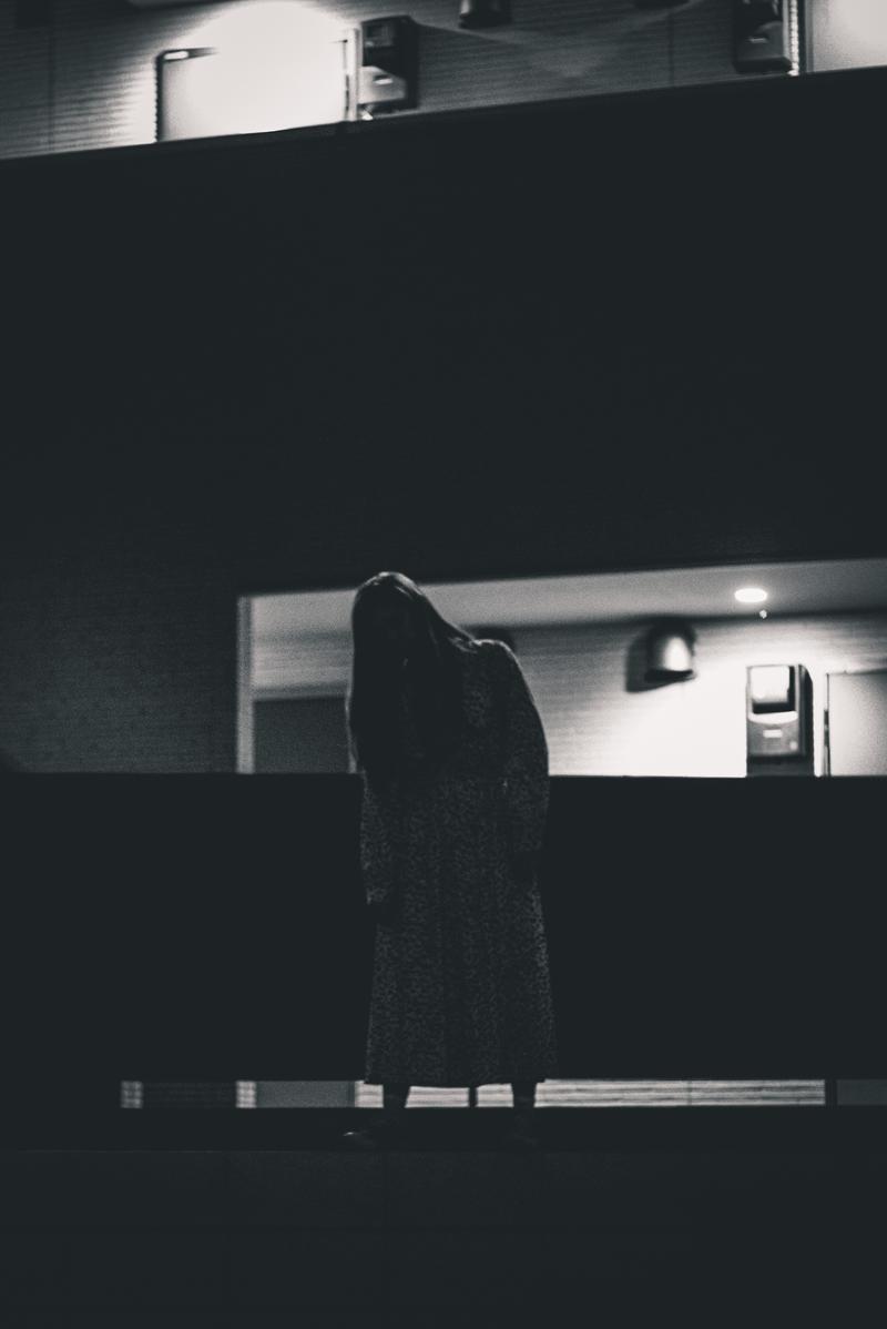 暗いマンション室内で窓の光に浮かぶ黒い衣装の女性の背中［モデル：ラミエル］