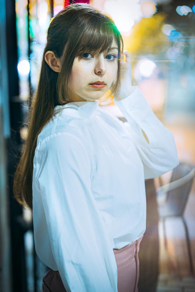 街灯りに包まれた夜景の中で振り返る白いシャツを着た女性のポートレート［モデル：ラミエル］