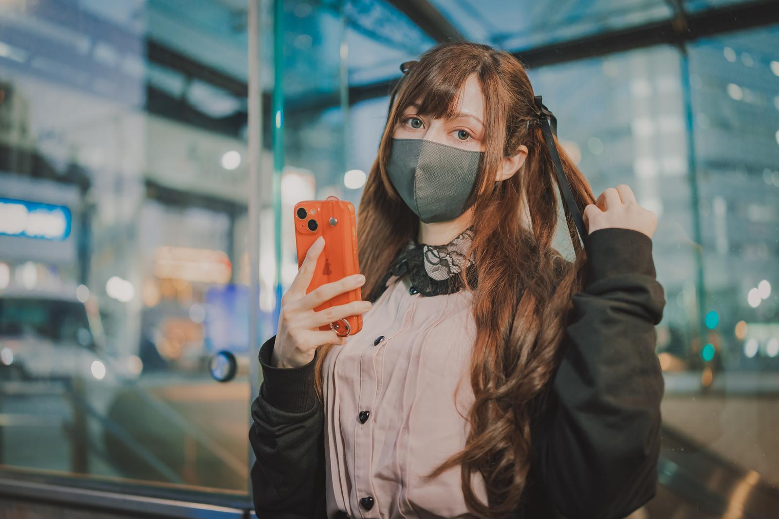黒マスクをした女性が赤いスマートフォンを持ち、夜の駅前で待ち合わせしている［モデル：ラミエル］
