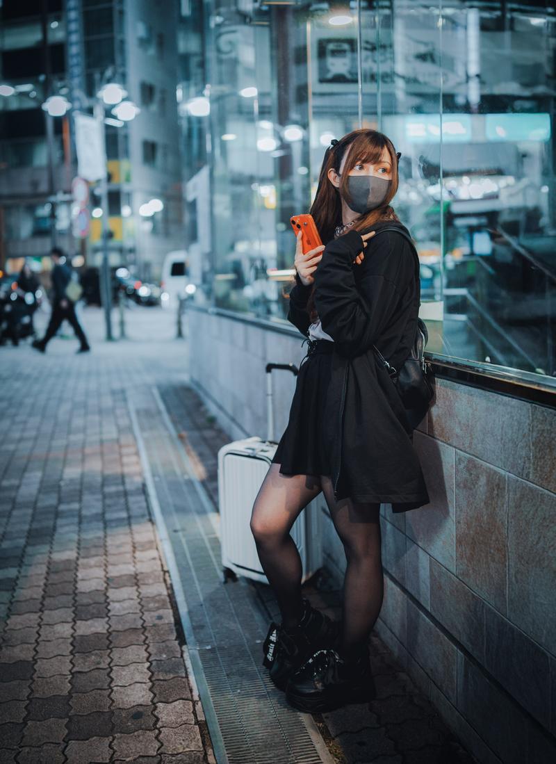 夜の駅前でキャリーケースを持つアメリカ人ハーフ女性がスマートフォンを見ている［モデル：ラミエル］