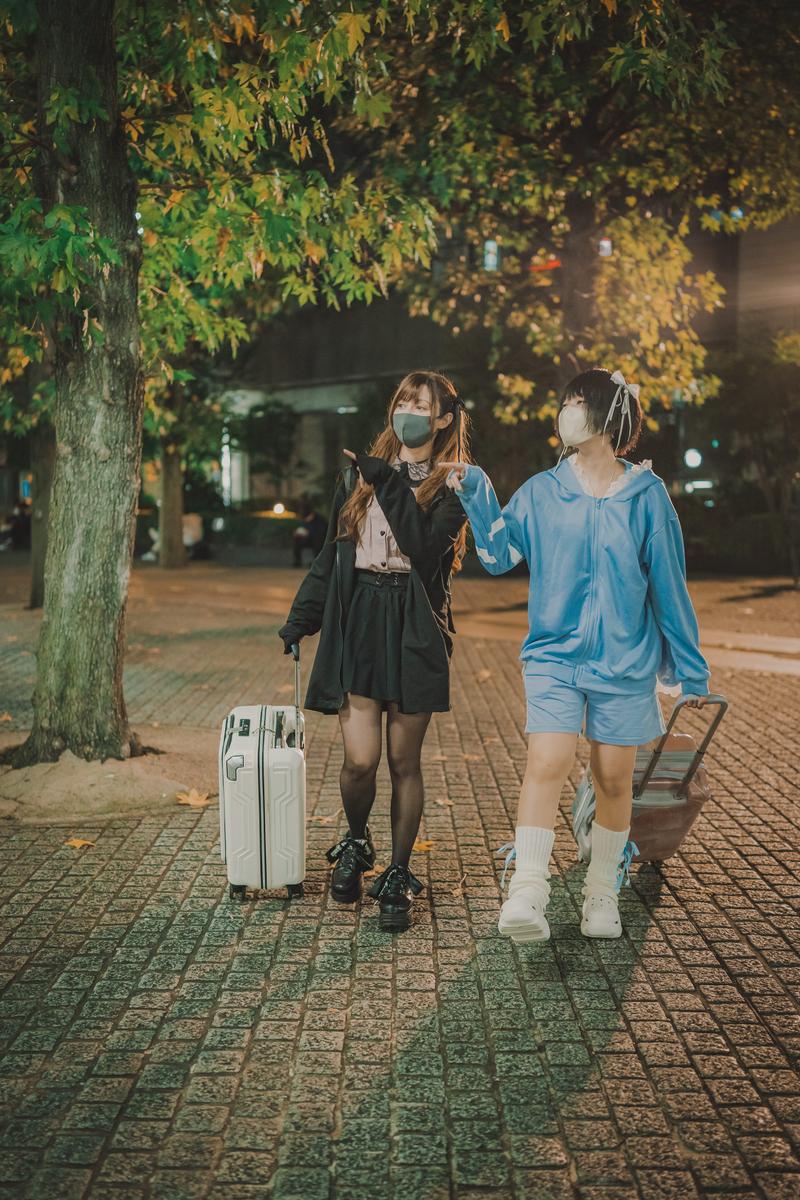 夜の街路樹の下で、マスク姿のふたりの女性がキャリーケースを引いて歩いている［モデル：ラミエル 緋真煉］
