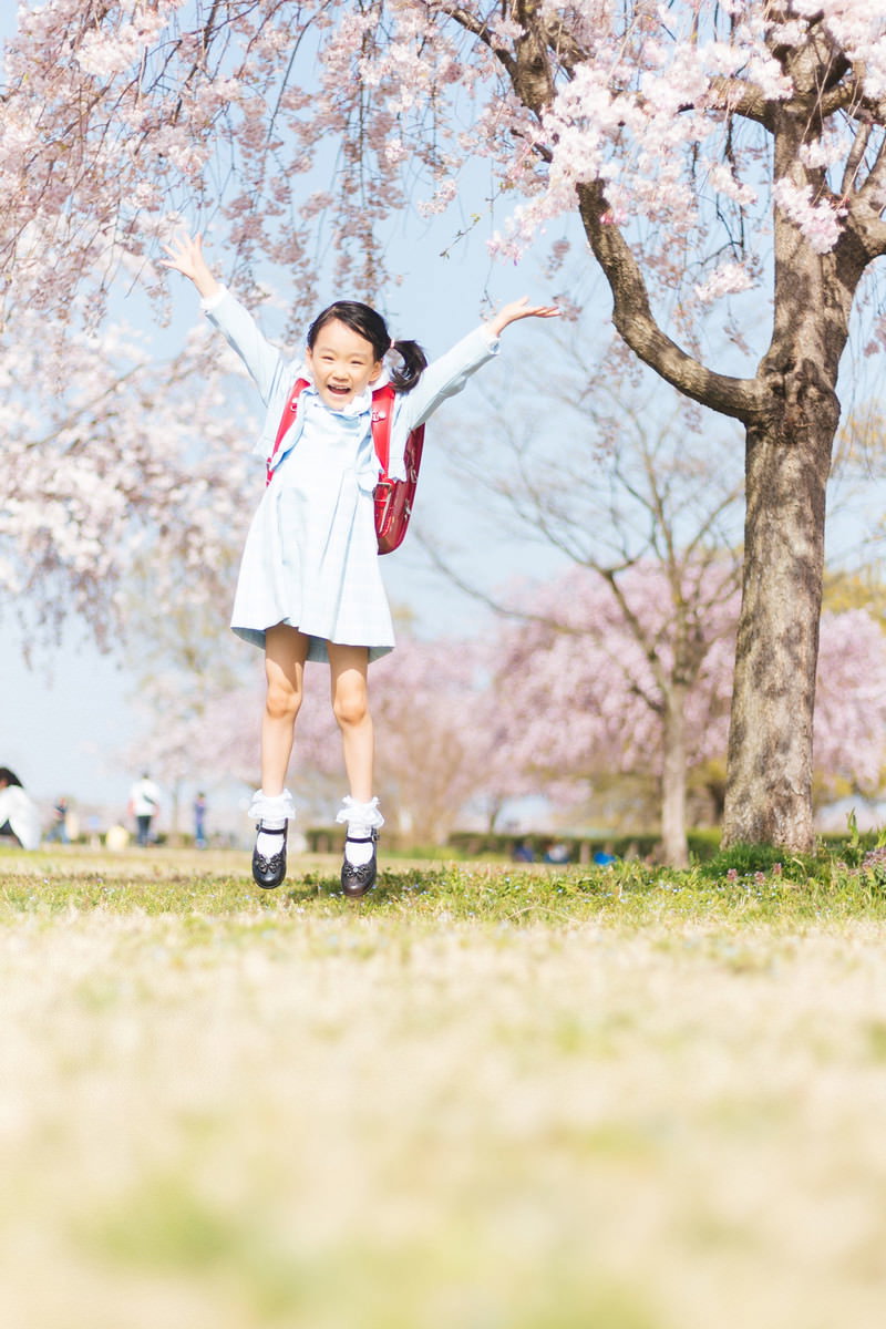 満開の桜の下で白いワンピースと赤いランドセルを身に着けた小学生が両腕を広げてジャンプしている［モデル：あんじゅ］