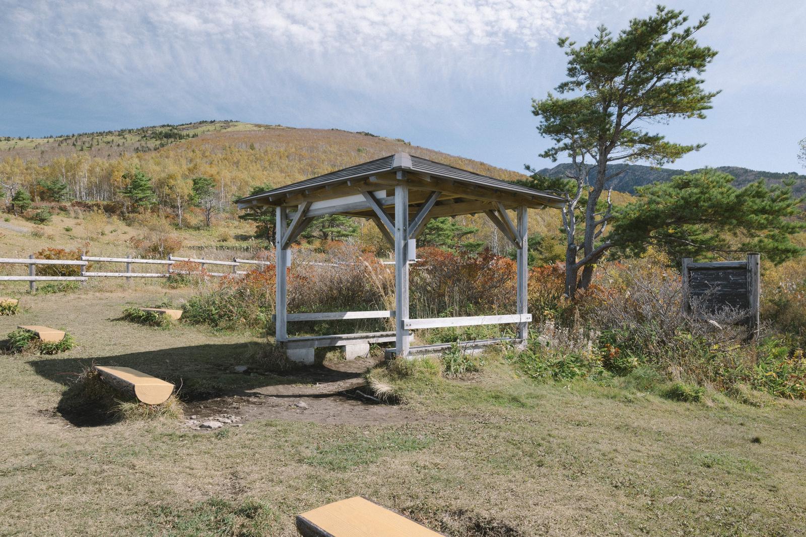 紅葉に囲まれた菅平牧場の木製東屋とベンチ、背景に秋の山々