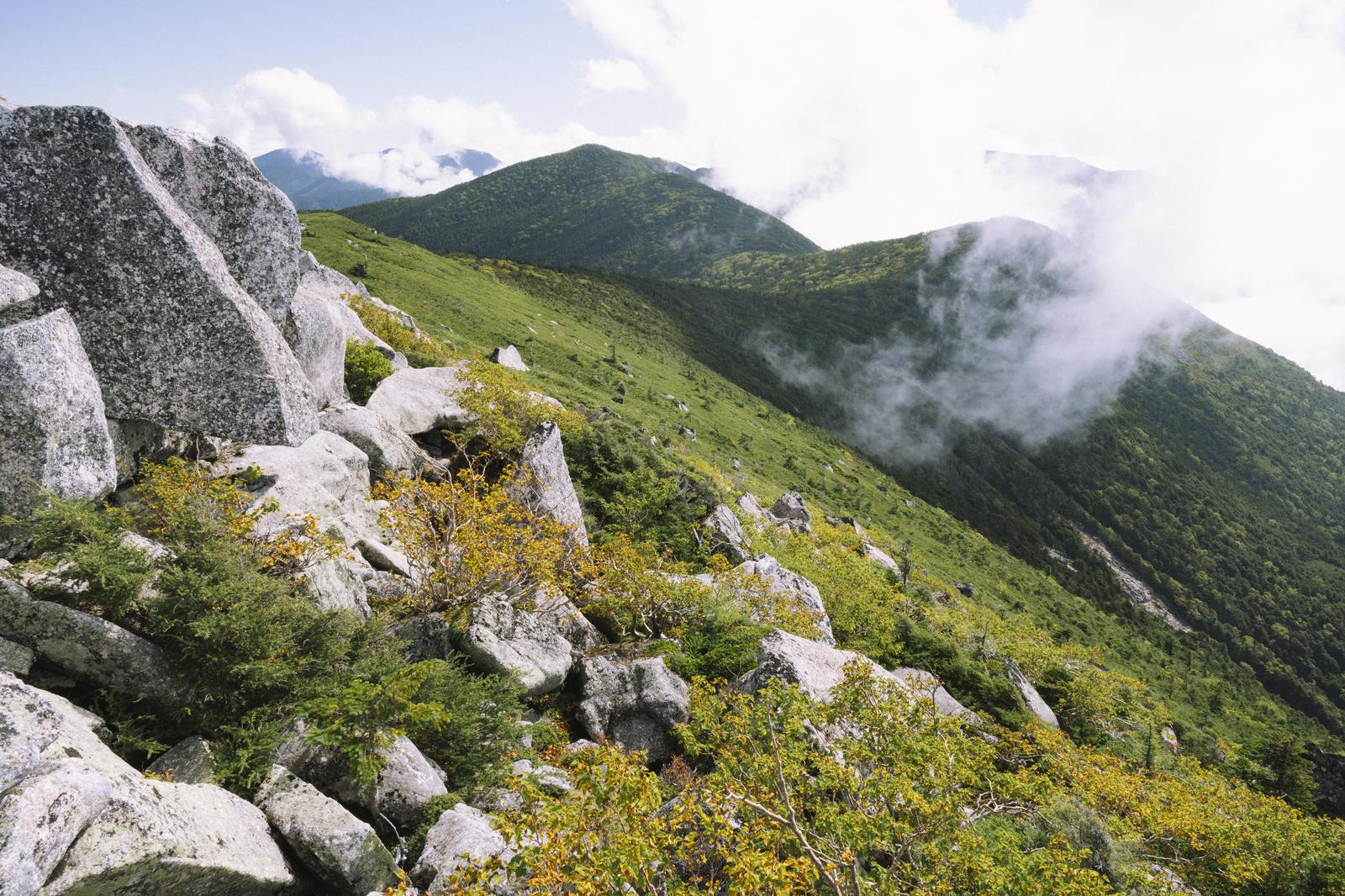 白い岩と緑の草地が続く金峰山の稜線から、雲がかかった山並みを望む風景