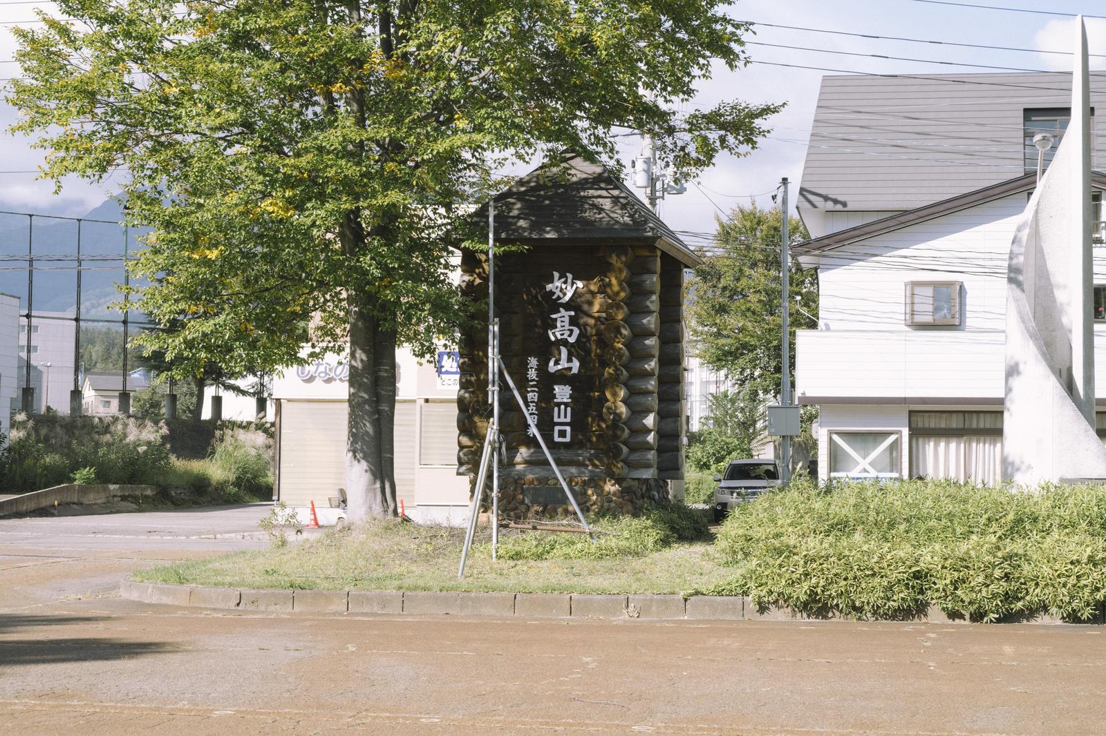 駅前に立つ妙高山登山口の標識と周囲の樹木や建物
