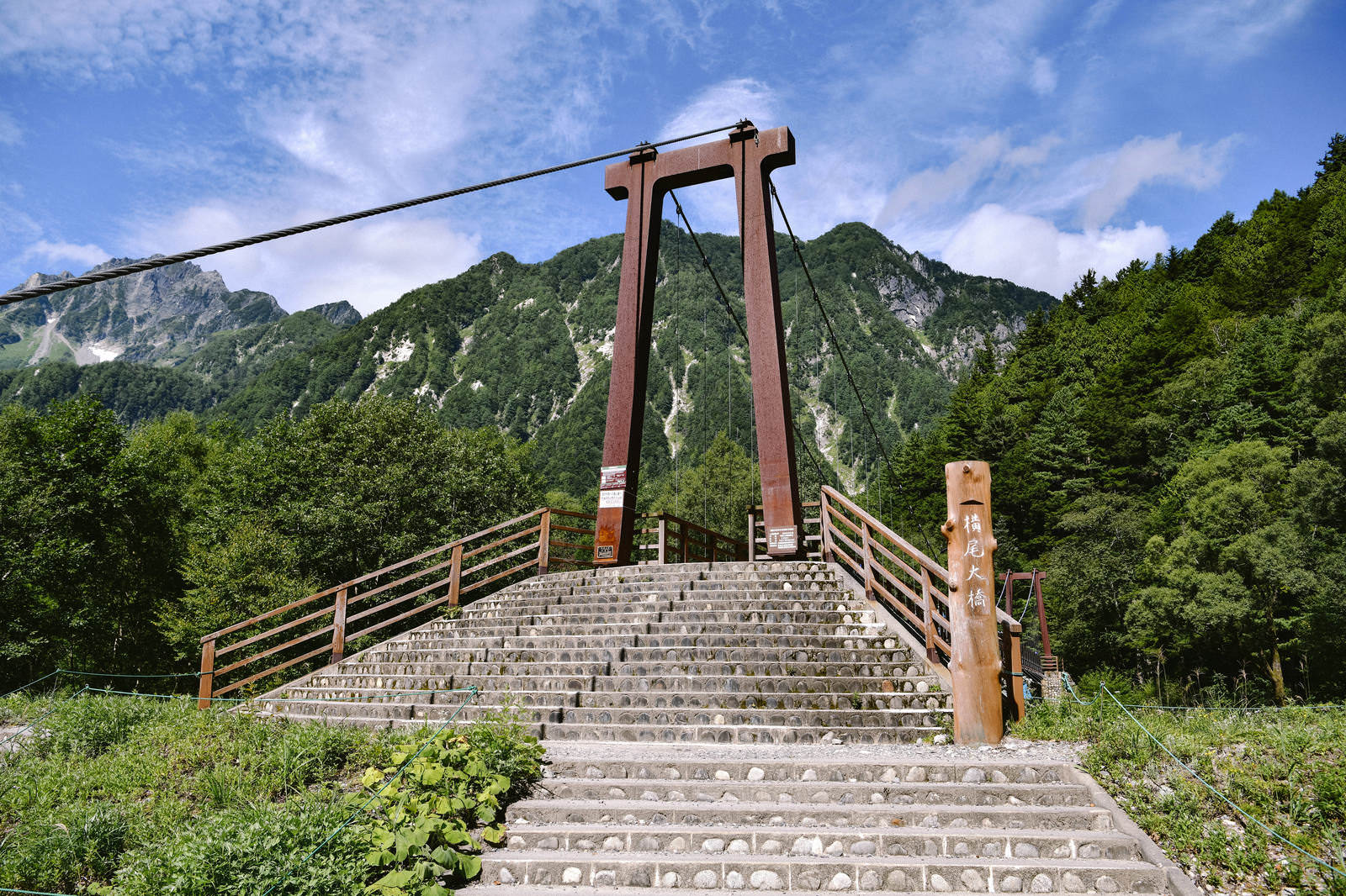 赤茶色の吊り橋の下から見上げた階段と橋の構造。背景に緑の山々