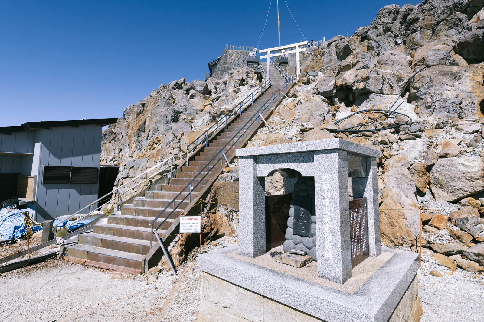 御嶽山の崖に建つ白い石造鳥居と奥の山小屋、急な木製階段