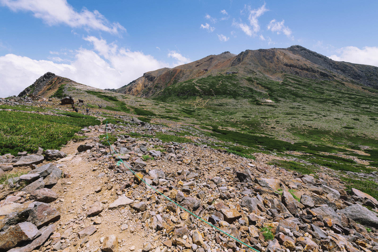 御嶽山の剣ヶ峰を望む登山道。手前に岩とロープ、背景に山肌と高山植物
