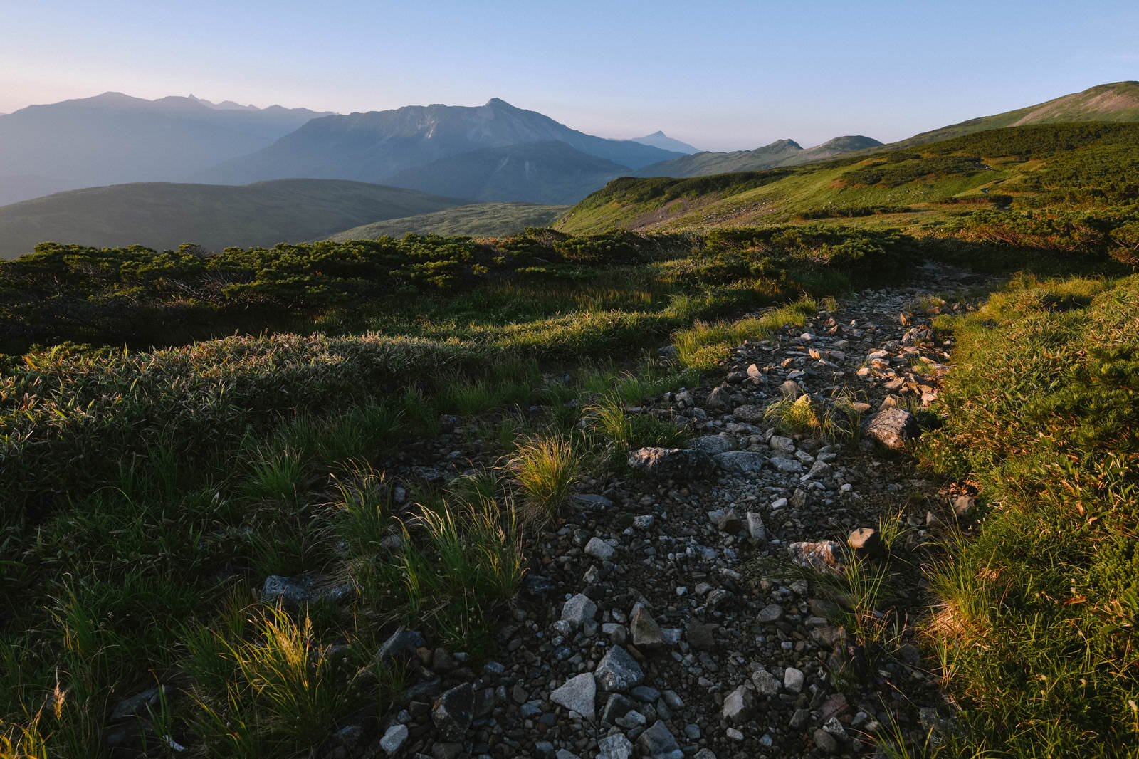 朝焼けの中、砂利道の登山道の先に黒部五郎岳の山並みを望む風景
