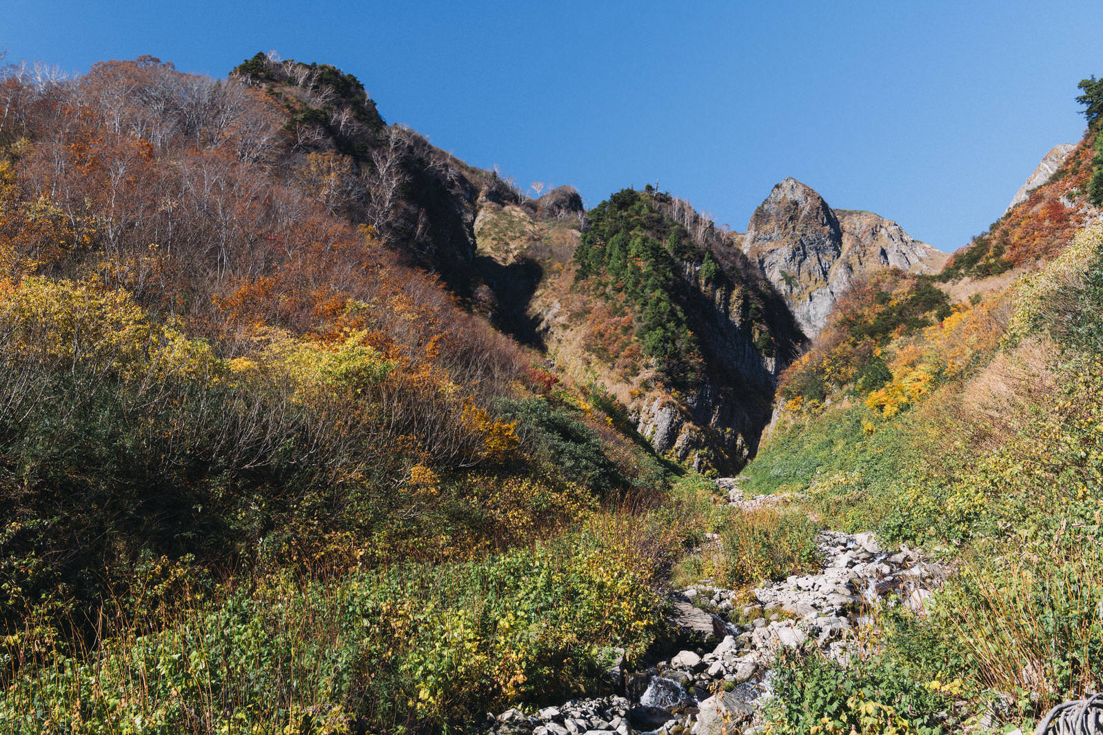 荒菅沢から見上げる秋の雨飾山、紅葉した山肌と岩場、青空が広がる