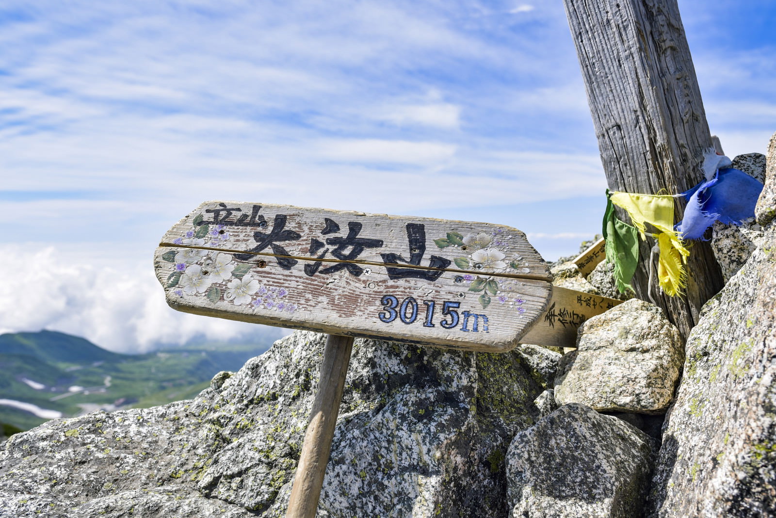 青空の下の岩場に立つ立山大汝山の木製山頂標と背景の山並み