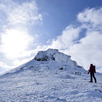 安達太良山の雪山山頂を眺める赤いウェアの登山者の写真