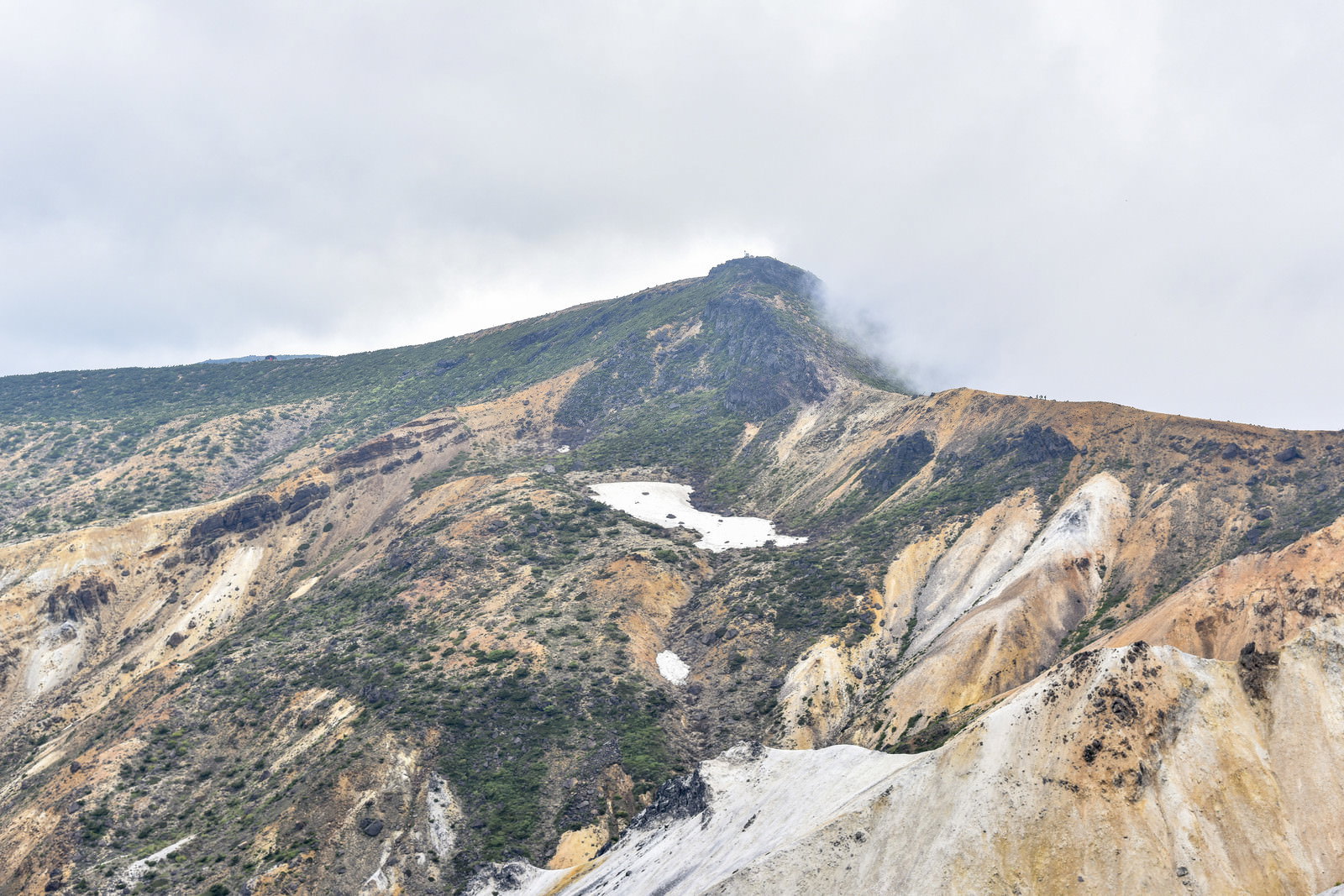 灰色の雲に覆われた安達太良山の山頂部分、荒々しい岩肌と残雪が見える