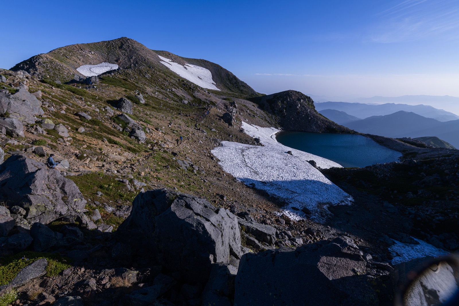 残雪が点在する岩場の登山道と青空、遠くの山並みが見える大汝峰への道中