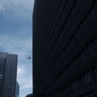 ビルの隙間の向こうへと跳んで行く飛行機のシルエットの写真