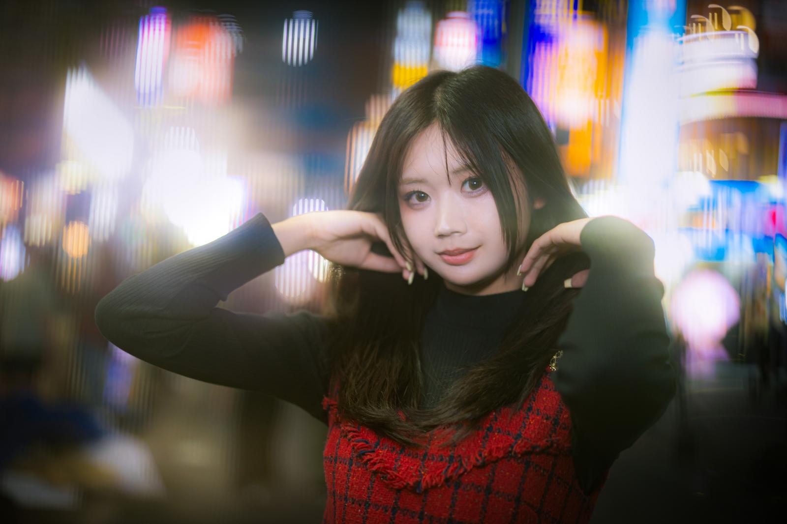 繁華街のネオン光を背景に、両手を頭の後ろに上げる黒シャツ姿の若い女性［モデル：れいか氏］