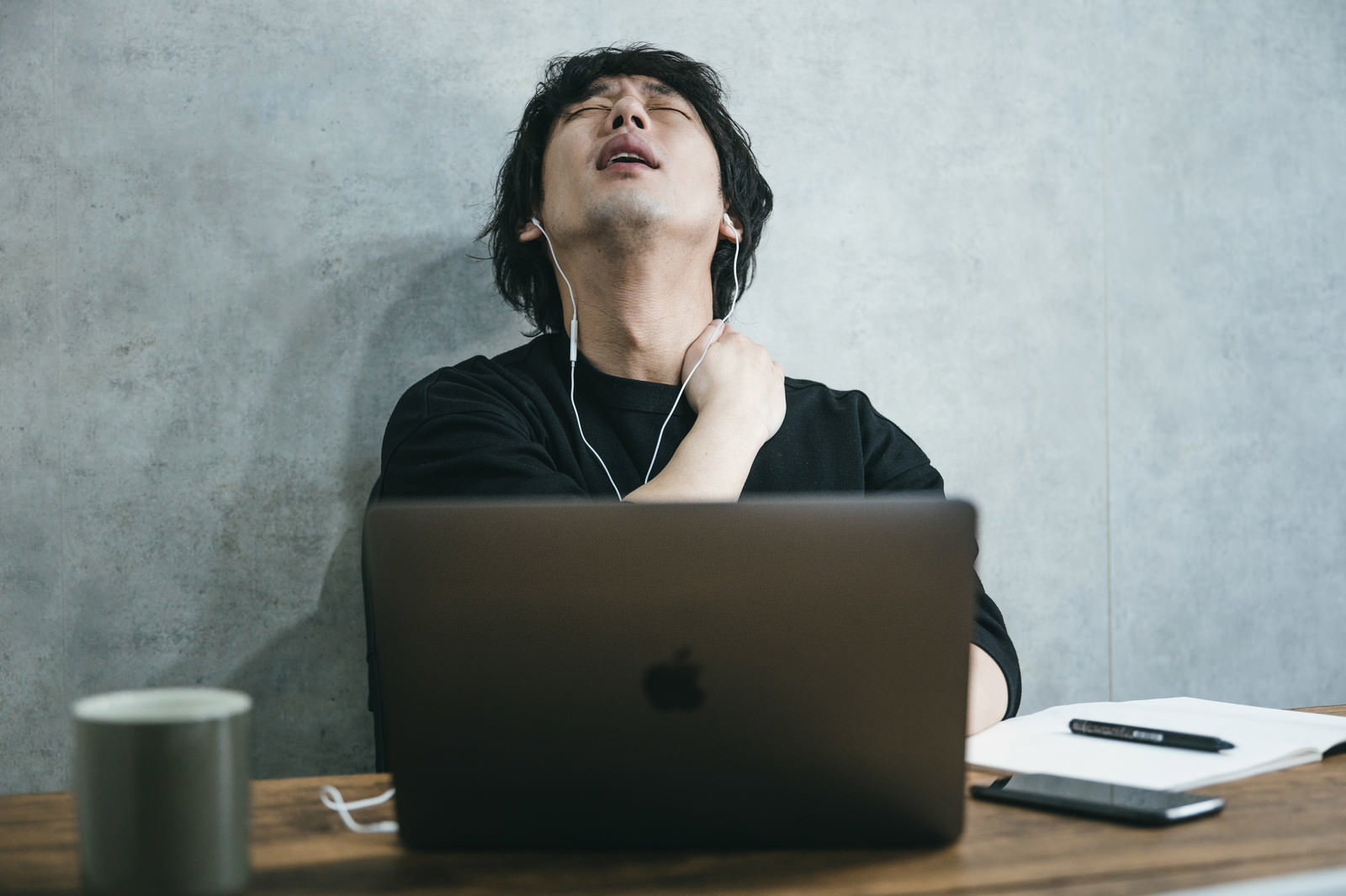 黒いシャツを着た男性がノートパソコンの前で疲れた表情で天井を見上げている［モデル：大川竜弥］