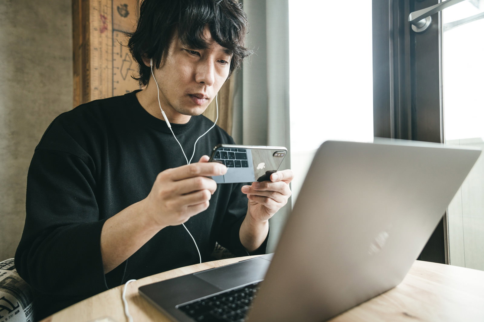ノートパソコンの前でスマートフォンのフロントカメラを使って身だしなみをチェックする黒いTシャツの男性［モデル：大川竜弥］