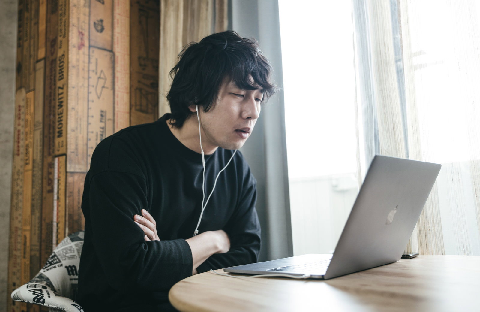 木製テーブルでノートパソコンに向かい腕を組んで考え込む黒い服の男性［モデル：大川竜弥］