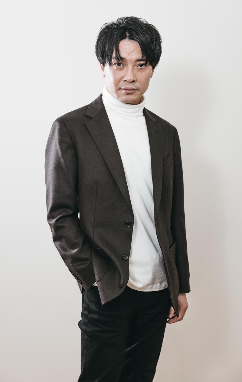 濃い茶色のジャケットと白いタートルネックを着た男性が正面を向いて立っている［モデル：金子周平］