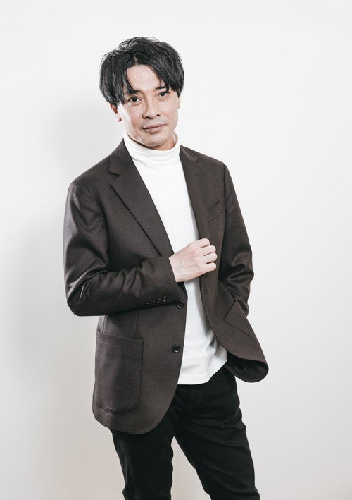 白いタートルネックにブラウンジャケットを羽織る取締役社長