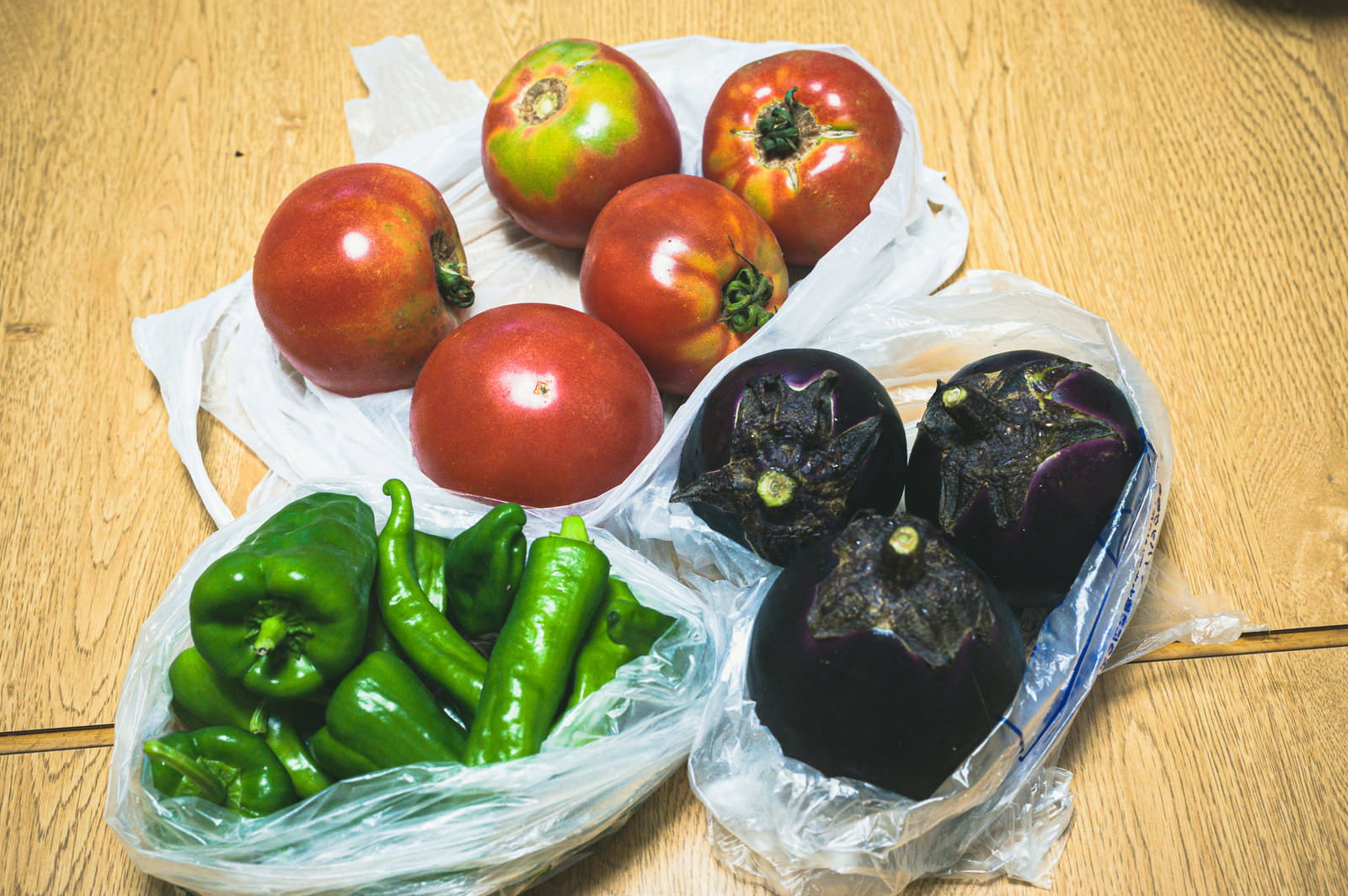 直売所で購入した新鮮な野菜。赤いトマト、緑のピーマン、紫色の茄子が袋に入って並んでいる