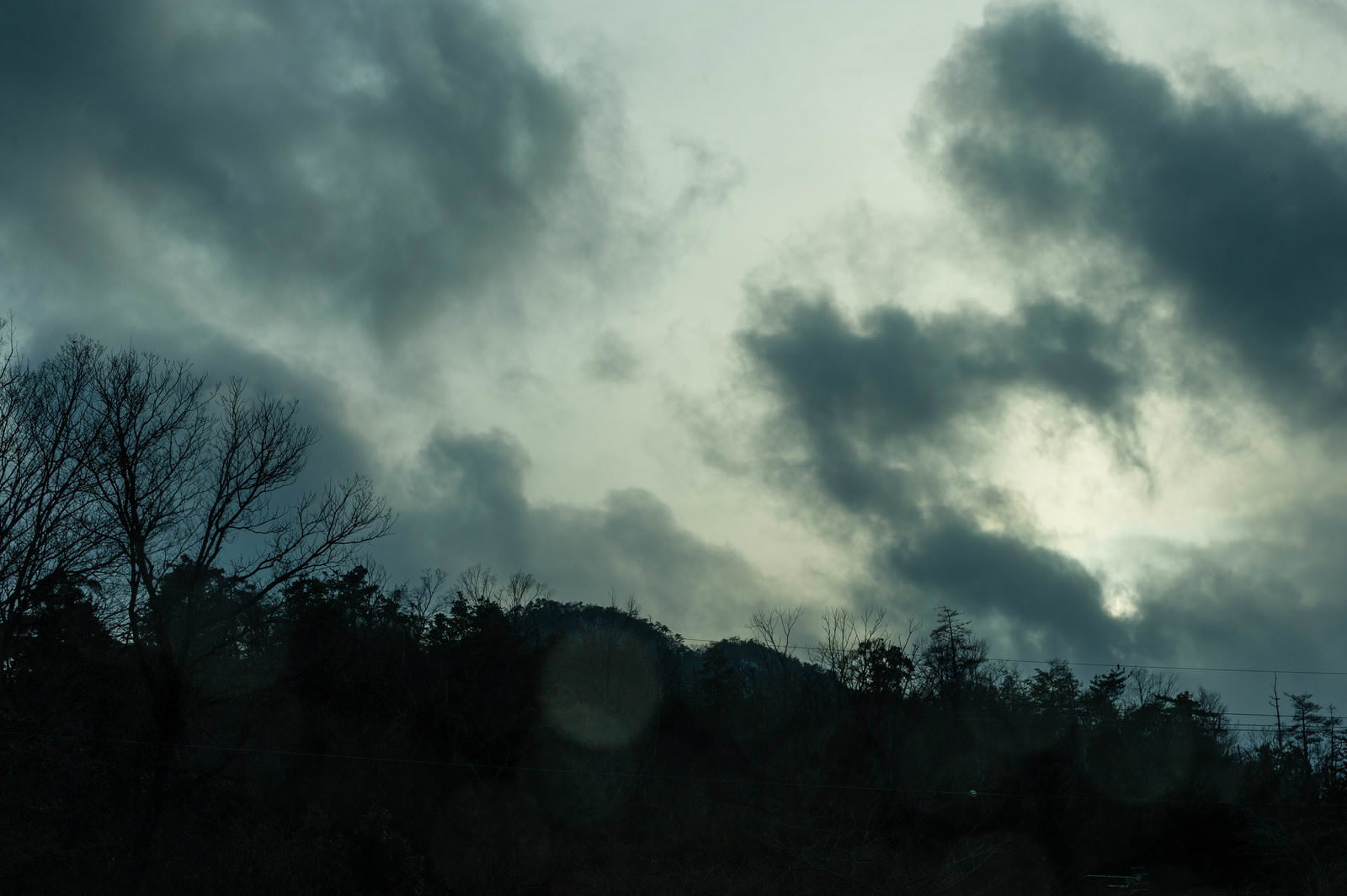 暗い雲が広がる空の下で、電線と木々のシルエットが見える風景