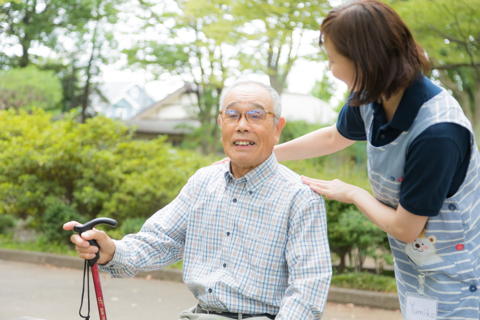 公園で車椅子に座るおじいさんと寄り添う介護士の女性［モデル：まーちゃん yumiko］