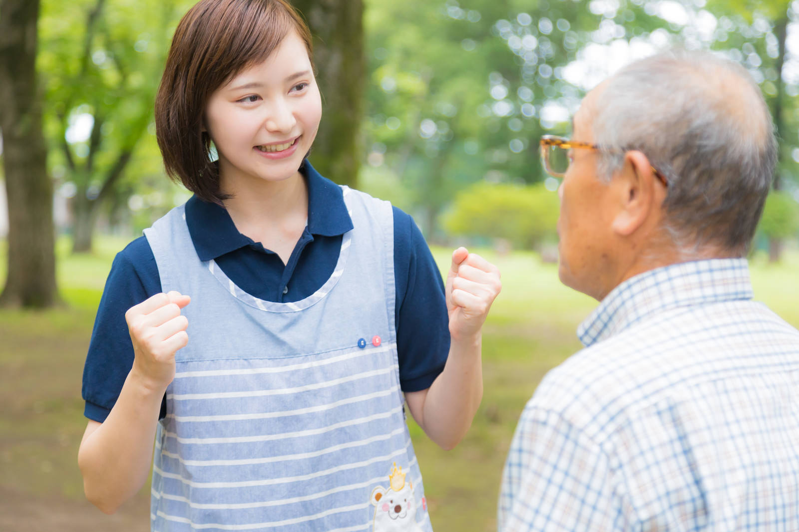 公園で高齢男性にガッツポーズで励ましの言葉をかける笑顔の女性介護士［モデル：まーちゃん yumiko］