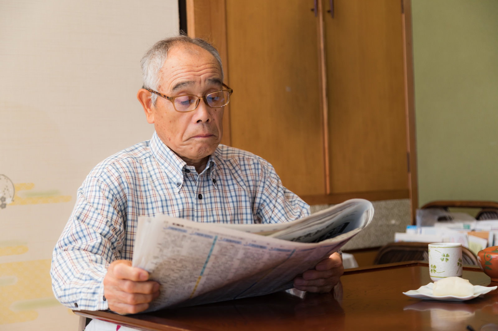 眼鏡をかけた高齢男性が新聞を読んでいる室内の様子［モデル：まーちゃん］