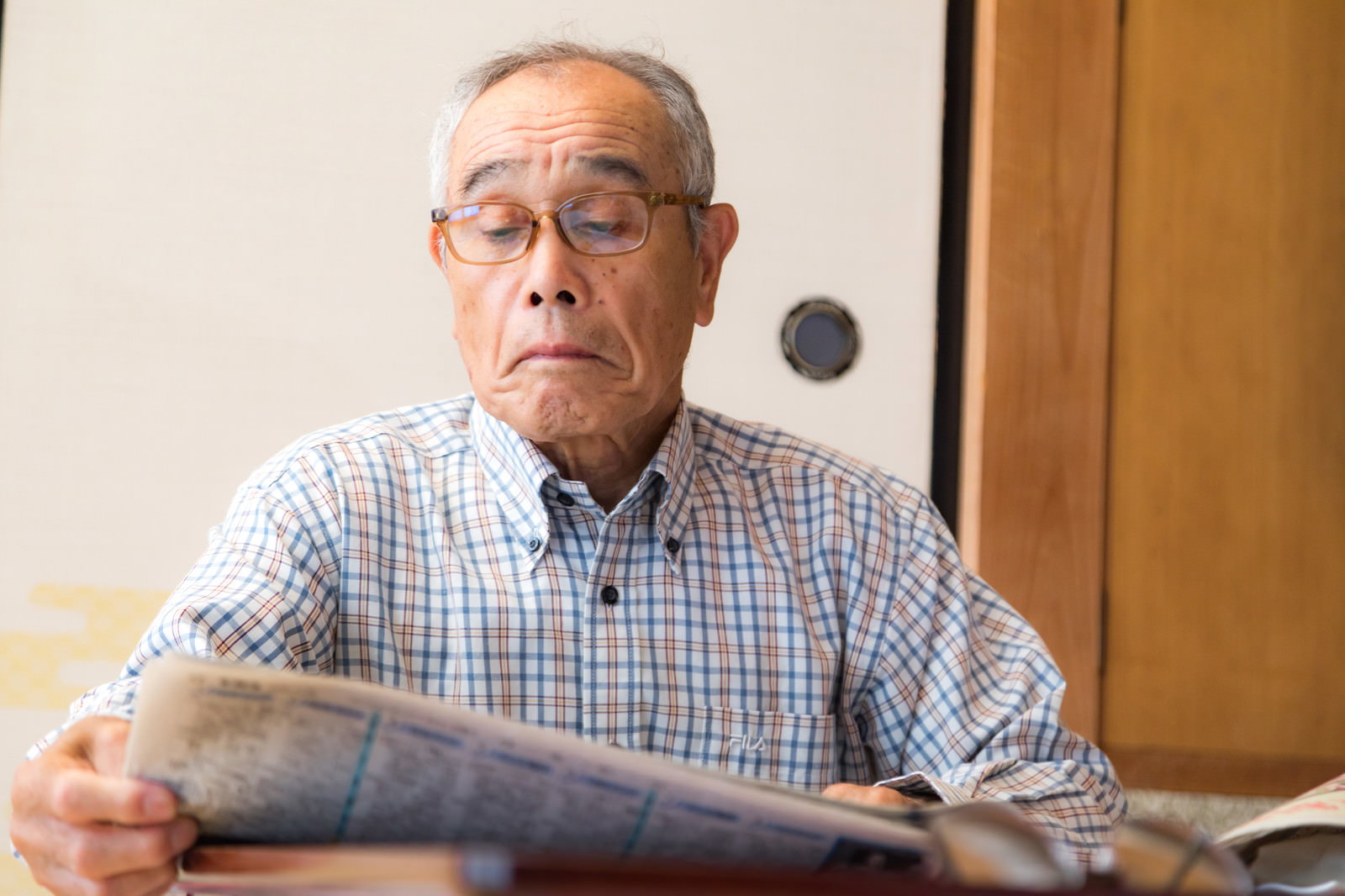 チェック柄のシャツを着た眼鏡のお爺さんが室内で新聞を読んでいる［モデル：まーちゃん］