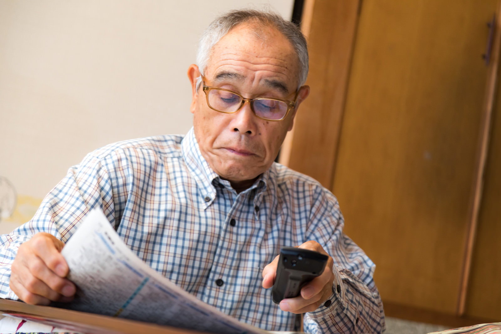チェックシャツを着た眼鏡のシニア男性が新聞を読みながらリモコンを持っている様子［モデル：まーちゃん］